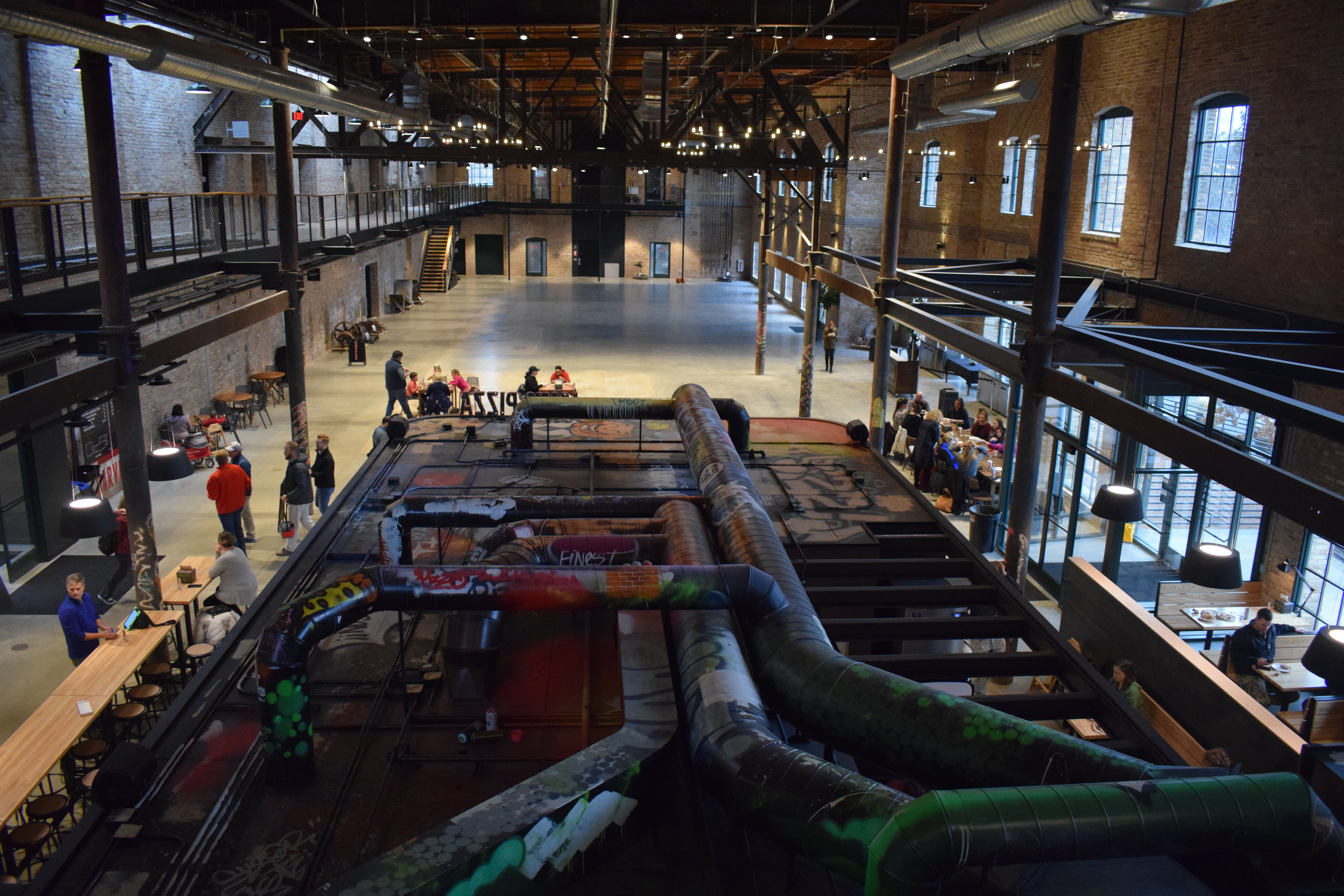 Exposed duct work with colorful graffiti is visible from the second floor inside the an industrial-looking building. Beyond the duct work, exposed beams, lighting, and a balcony can be seen, as well as a large open space on the first floor with passersby.  