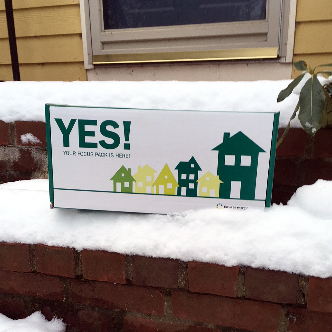 A white box with the word "YES!" in green capital letters is laid on a snowy step on a brick staircase in front of a door. There are graphic images of houses on the box in yellow and green colors.