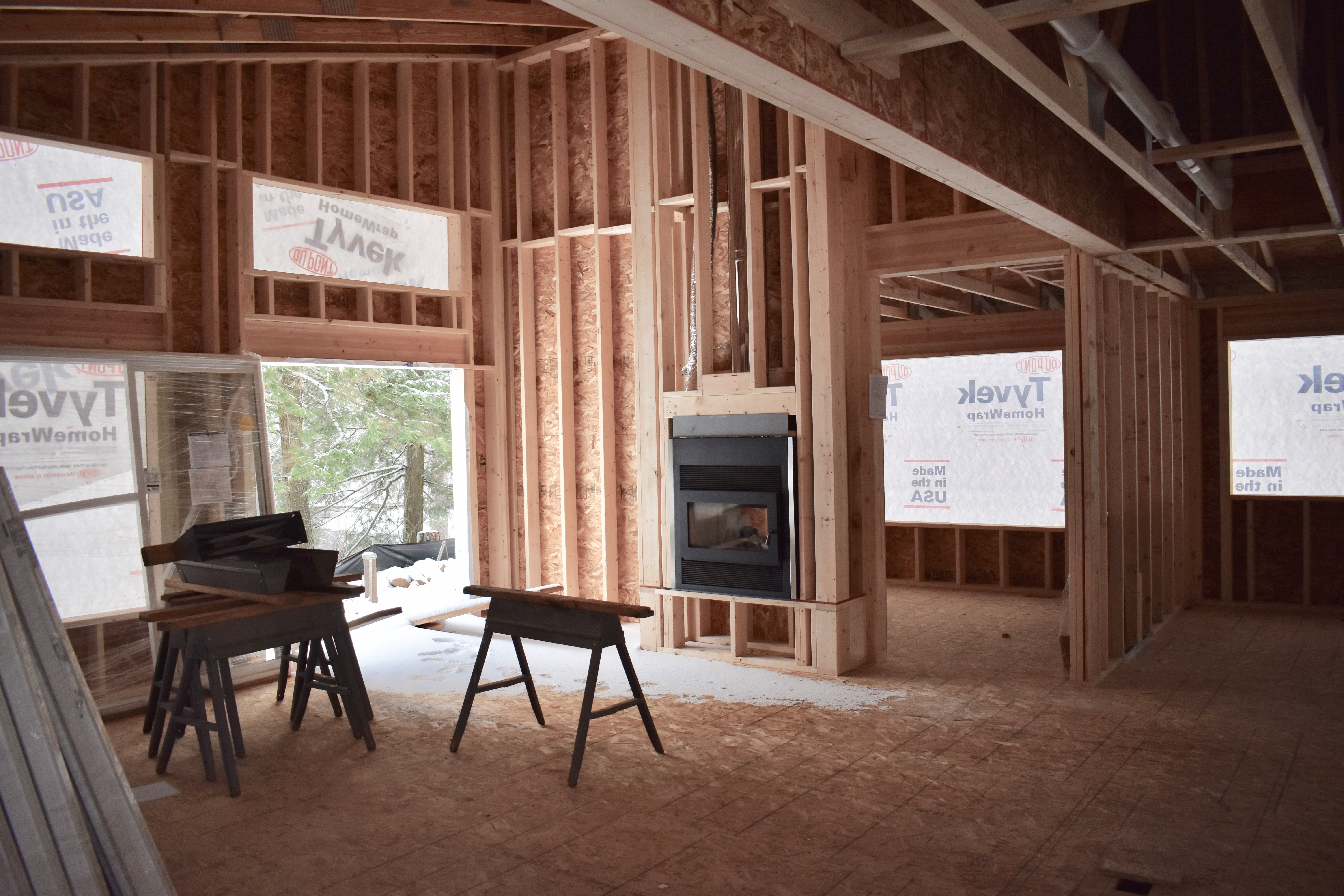 Interior of a home currently under construction - exposed beams, unfinished walls, and unfinished flooring can be seen. There is a fireplace installed in the wall. 