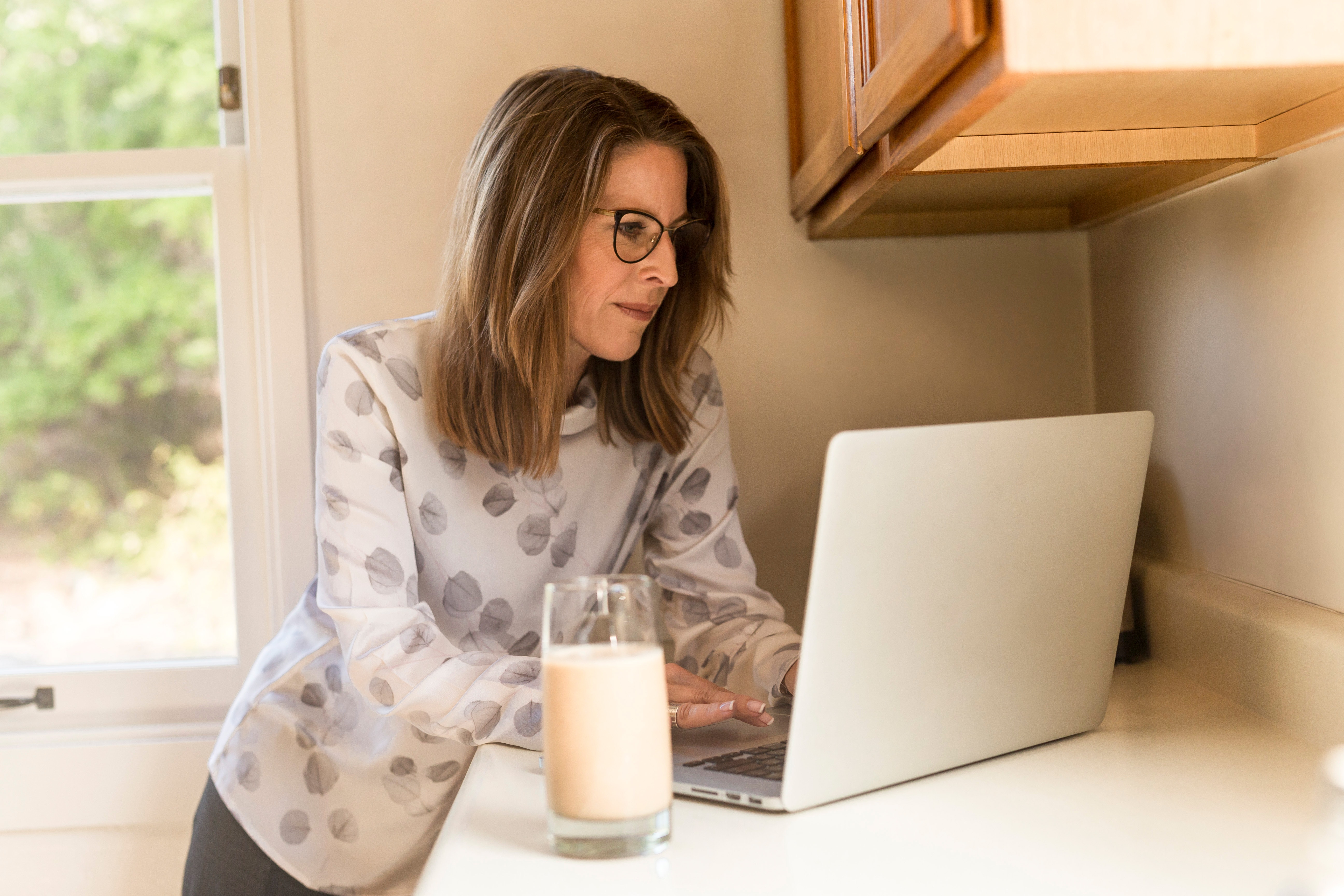 Woman wearing glasses stands at a kitchen counter while using a laptop. There is a glass of milk on the counter.
