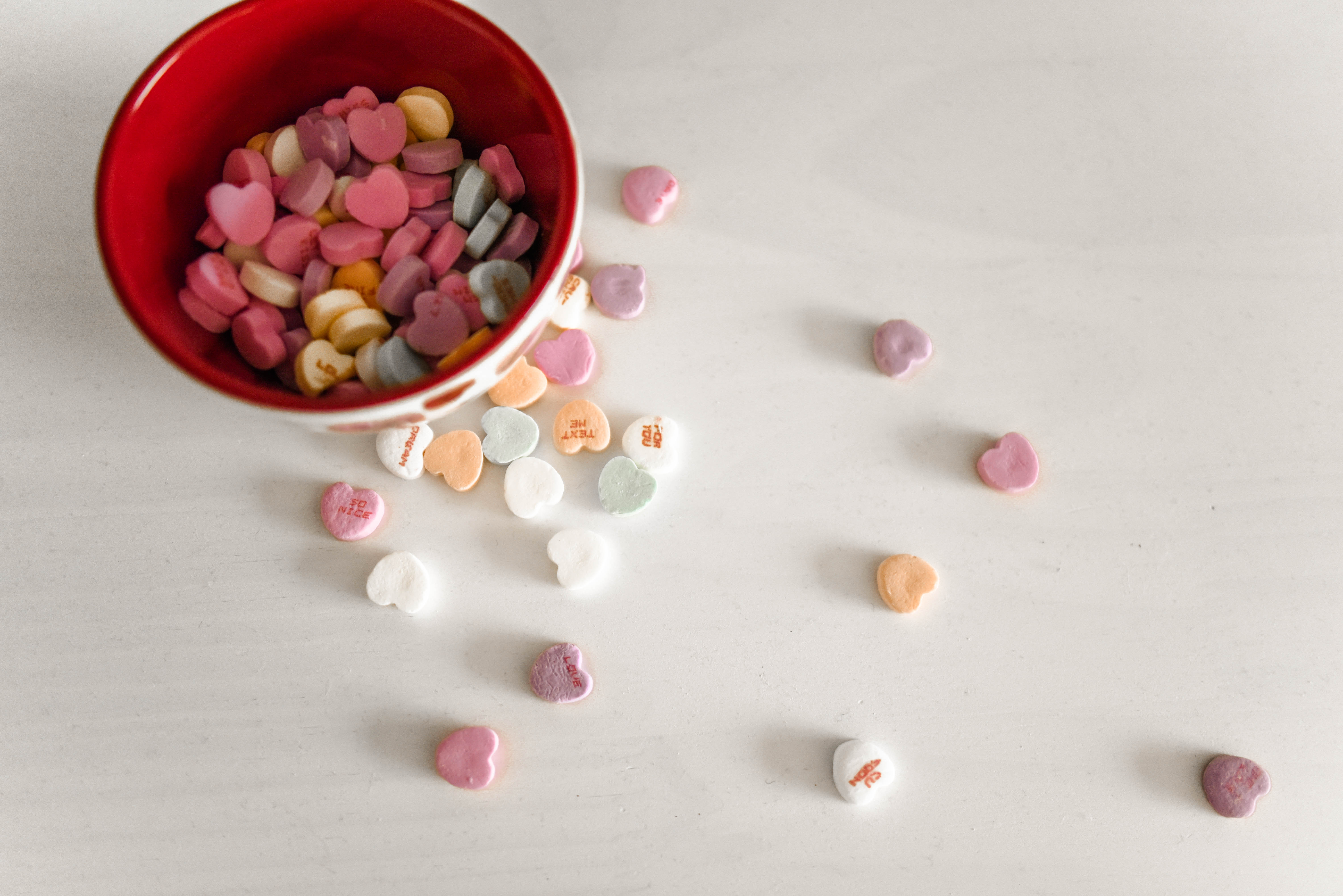 Red bowl of colorful conversation hearts on a white backdrop with conversation heart candy spilled over the side.